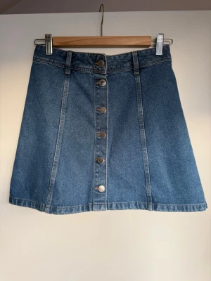 Blå jeanskjol med knappar från Boomerang - Säljer en klassisk blå jeanskjol från H&M med A-linjeform och knappar framtill. Kjolen har bälteshällor och sömdetaljer som ger en snygg retrovibe. Perfekt att matcha med sneakers eller boots.