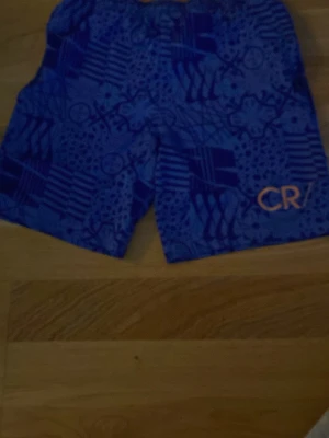 Blå mönstrade shorts CR - Snygga blå shorts med unikt grafiskt mönster och CR-logga i orange på benet. Perfekta för sommaren och har en avslappnad passform. Tillverkade i ett lätt och bekvämt material.