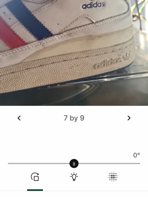 Adidas vita sneakers med röd/blå detalj - Klassiska vita sneakers från Adidas med högt skaft och ikoniska ränder i rött och blått på sidan. Skorna har perforerad tå, vit sula med Adidas-logga och blå yttersula. Tillverkade i läder med textildetaljer och snörning framtill.
