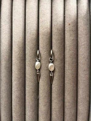 Silvriga örhängen med pärldetalj - Säljer ett par stilrena örhängen i silverfärg med oval vit pärldetalj och en spetsig konformad hängande dekoration. Örhängena har en modern och edgy look, perfekta för att lyfta din outfit. Materialet ser ut att vara metall och pärlimitation.