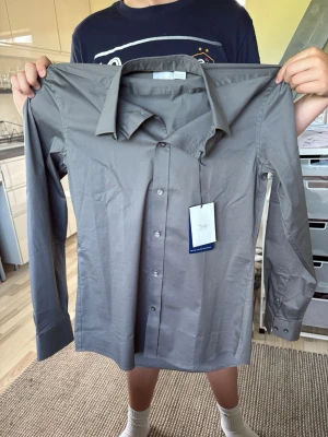 Grå skjorta från Tiger of Sweden - Snygg grå skjorta från Tiger of Sweden i slim fit-modell. Skjortan har klassisk krage, långa ärmar och knäppning framtill. Tillverkad i bomull för en skön känsla och stilren look. Perfekt för dig som gillar minimalistisk och clean stil.