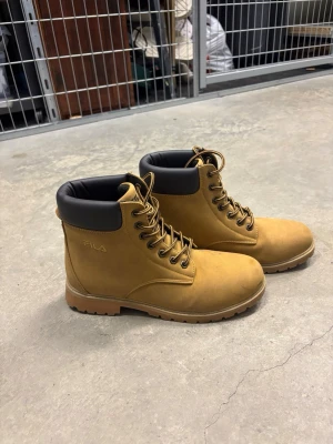 Bruna kängor från Fila  - Snygga bruna kängor från Fila med robust sula och svart vadderad kant upptill. Kängorna har snörning och är tillverkade i mockaliknande material. Perfekta för dig som vill ha en klassisk och stilren look till hösten eller vintern.