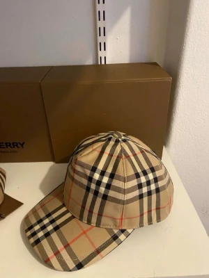 Burberry beige rutmönstrad keps - Säljer en klassisk keps från Burberry i beige med det ikoniska rutmönstret i svart, vitt och rött. Kepsen har böjd skärm och justerbart spänne bak. Tillverkad i bomull för en skön och stilren look.