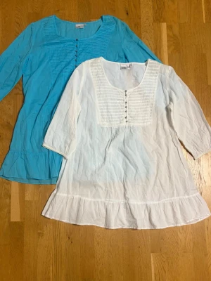 Två tunna blusar  - Säljer två luftiga blusar, en i vitt och en i turkos. Båda har trekvartsärm, knappar framtill och dekorativa sömmar vid bysten. Perfekta till jeans eller shorts. Materialet känns lätt och skönt mot huden.
