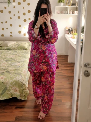 Blommig pyjamas i rosa och lila - Säljer en färgglad pyjamas med blommigt mönster i rosa, lila och orange toner. Setet består av en långärmad överdel med lös passform och matchande vida byxor. Perfekt för mysiga hemmakvällar. Materialet känns mjukt och luftigt.