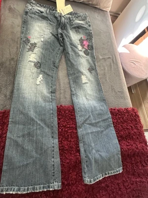 Bootcut jeans med broderi och slitningar - Snygga ljusblå bootcut jeans med slitningar och broderade detaljer i svart och rosa på framsidan. Jeansen har klassisk femficksmodell och är tillverkade i denim. Perfekta för dig som gillar en avslappnad men ändå unik stil.
