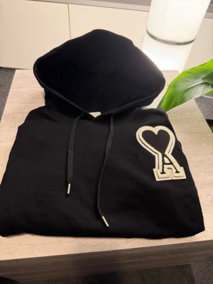 Svart Ami Paris hoodie - Säljer en svart hoodie från Ami Paris med den ikoniska vita hjärtformade A-loggan på bröstet. Perfekt för en avslappnad och trendig stil. Materialet är mjukt bomullstyg.