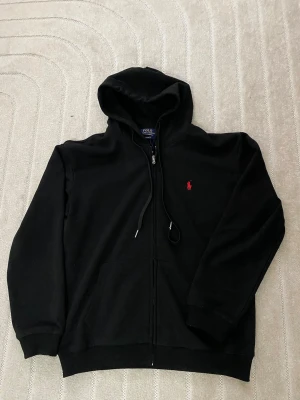 Svart hoodie från Polo Ralph Lauren - Svart hoodie med dragkedja från Polo Ralph Lauren. Klassisk design med huva och dragsnören, två fickor framtill och den ikoniska röda loggan broderad på bröstet. Tillverkad i mjukt bomullsmaterial som är skönt att ha på sig.