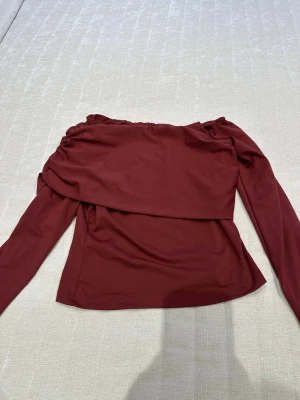Vinröd offshoulder topp - Trendig vinröd offshoulder topp med långa ärmar och snygg drapering över bröstet. Perfekt för dig som vill ha en unik och stilren look. Tillverkad i mjukt syntetmaterial som sitter skönt på kroppen.