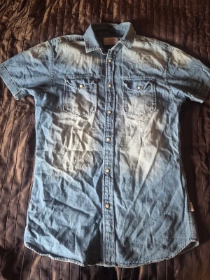Blå jeansskjorta med kort ärm - Snygg blå jeansskjorta i bomull med korta ärmar och tryckknappar framtill. Skjortan har två bröstfickor med lock och knappdetaljer. Perfekt för en avslappnad look