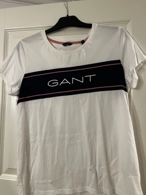 Vit GANT t-shirt med logga - Vit t-shirt från GANT med korta ärmar och rund halsringning. Framtill finns en bred marinblå rand med GANT-logga i vitt och tunna rosa linjer ovan och under. Perfekt för en stilren och sportig look.