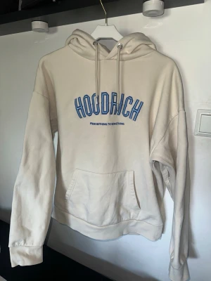 Beige hoodie från Hoodrich - Snygg beige hoodie från Hoodrich med blå text på bröstet där det står 'HOODRICH' och 'FROM NOTHING TO SOMETHING' under. Hoodien har stor känguruficka framtill, dragsko i huvan och ribbade muddar vid ärmar och nederkant. Även en gömd ficka på ena armen. Perfekt för en chill och avslappnad stil.