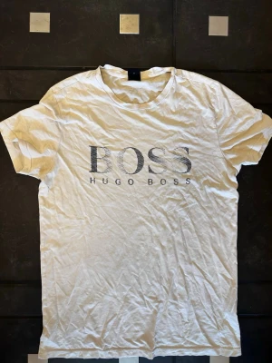 Vit t-shirt från Hugo Boss - Vit t-shirt från Hugo Boss med stort BOSS-tryck på bröstet. Klassisk rund hals och korta ärmar. Tillverkad i mjuk bomull och har en clean, stilren look som passar perfekt till jeans eller shorts.