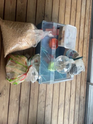Hamsterbur med tillbehör och spån - Säljer ett komplett hamsterkit med genomskinlig bur, plastmat- och vattenskålar, springboll, transportbur, matskopa, samt flera påsar spån och hamsterfoder. Perfekt för dig som vill starta med hamster! Allt i plast och metall, färgerna är mestadels transparent, orange och grå.