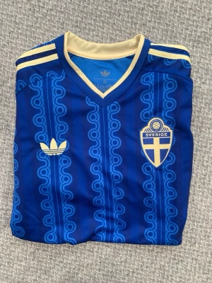 Blå Sverige Adidas fotbollströja - Säljer en blå fotbollströja från Adidas med Sveriges landslagsemblem på bröstet. Tröjan har retroinspirerat mönster, V-ringning och gula detaljer på krage och axlar. Tillverkad i lätt och ventilerande material, perfekt för match eller träning.