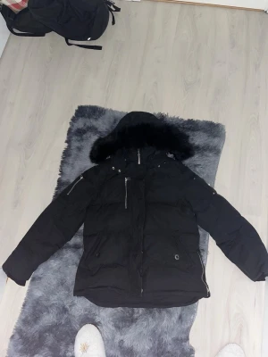 Svart dunjacka med pälshuva Moose Knuckles - Säljer en svart dunjacka från Moose Knuckles med stor pälskantad huva och flera dragkedjefickor. Jackan har den ikoniska metall-loggan på ärmen och är perfekt för kalla vinterdagar. Stilig och rejäl design som håller dig varm hela säsongen.