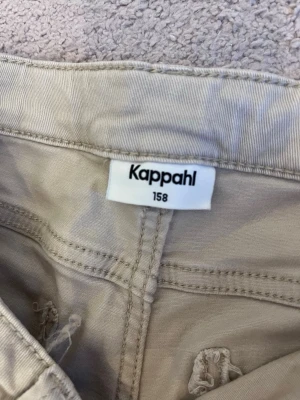 Beige cargobyxor från Kappahl - Säljer ett par beige cargobyxor från Kappahl i storlek 158. Byxorna har raka ben, stora fickor på sidorna och bak, samt bälteshällor. Tillverkade i ett mjukt bomullstyg som är skönt att ha på sig. Perfekta för en avslappnad och trendig look.