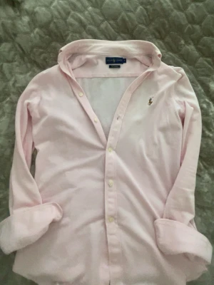 Ljusrosa skjorta från Ralph Lauren - Säljer en klassisk ljusrosa skjorta från Ralph Lauren med den ikoniska broderade loggan på bröstet. Skjortan har lång ärm, knappar framtill och är tillverkad i mjuk bomull. Perfekt för en clean och stilren look. Perfekt till sommaren⭐️☀️