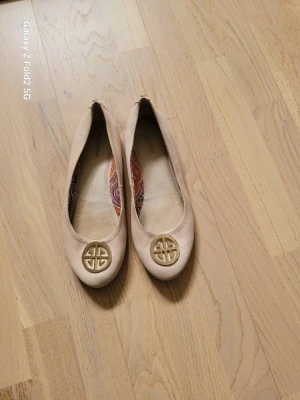 Beige ballerinaskor från Depeche - Snygga beige ballerinaskor från Depeche med rund tå och guldfärgad metallogo framtill. 