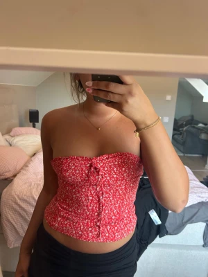 Röd blommig bandeau tubtopp - Säljer en röd bandeau tubtopp med små vita blommor och snörning framtill. Toppen är axelbandslös och croppad, perfekt för sommaren. Materialet känns lätt och bekvämt, troligen bomull eller polyester. Snygg att styla med både jeans och kjol. 