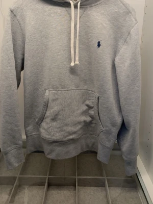 Ljusgrå hoodie från Polo Ralph Lauren - Ljusgrå hoodie från Polo Ralph Lauren med klassisk känguruficka och vit dragsko i huvan. Broderad blå logga på bröstet. Tillverkad i mjuk bomullsblandning för en chill och bekväm stil. Perfekt för dig som gillar enkel och stilren design.