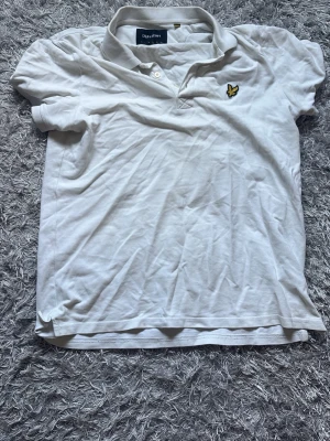 Vit pikétröja från Lyle & Scott - Klassisk vit pikétröja från Lyle & Scott med korta ärmar och gul logga på bröstet. Tröjan har krage och knappar vid halsen, tillverkad i mjuk bomull för en bekväm känsla. OBS notera små små hål i armhålan. Det står storlek L men skulle säga den är storlek M