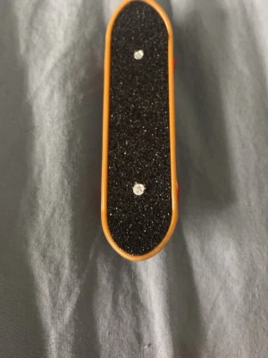 Fingerboard med högtalardesign - Säljer en fingerboard med svart grepptejp och coolt motiv av högtalare i lila, rosa och vitt på undersidan. Brädan har röda hjul och silverfärgade truckar. Perfekt för dig som gillar att göra tricks med fingrarna!