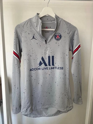 Grå PSG träningströja långärmad - Snygg grå långärmad träningströja från PSG i samarbete med Jordan. Tröjan har blå logga och text, halv dragkedja, samt röda, vita och blå detaljer på ärmarna. Materialet är funktionspolyester och har ett stänkmönster över hela plagget.