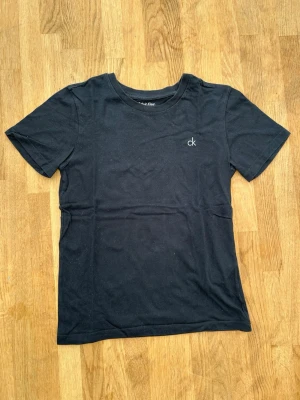 Svart t-shirt från Calvin Klein - En stilren svart t-shirt från Calvin Klein med diskret 'ck'-logga broderad på bröstet. Klassisk passform med rund halsringning och korta ärmar. Perfekt basplagg i mjuk bomull som funkar till allt.