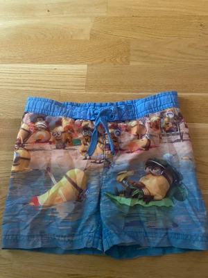 Blå Minions badbyxor med tryck - Säljer ett par blå badbyxor med färgglatt Minions-tryck. De har snörning i midjan och elastisk linning för skön passform. Perfekta för stranden eller poolen och har ett lekfullt motiv med Minions på semester. Materialet känns som snabbtorkande polyester.