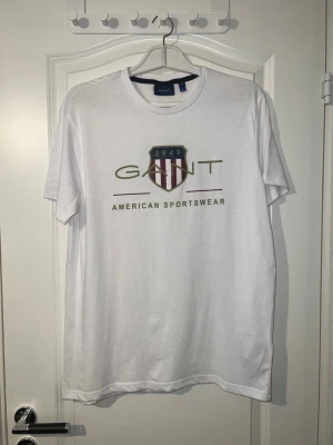 GANT t-shirt  - Vit t-shirt från GANT med rund hals och tryck framtill. Trycket består av GANT-loggan, ett sköldemblem med rödvita ränder och texten 'American Sportswear'. Klassisk och stilren design, perfekt till jeans eller shorts. Finns att hämta i Södertälje.