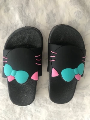 Hello Kitty Barn sandaler med rosett - Två par sandaler med Hello Kitty-motiv. Ena paret är svart med turkos och rosa rosett, det andra är vitt med röd rosett. Båda har öppna tår och bred rem över foten. Tillverkade i syntetmaterial och har en bekväm, platt sula. Stlk kvar 24,25,26,28 och 30 