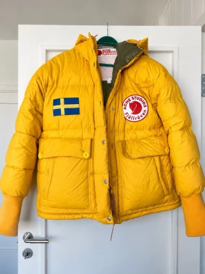 Gul dunjacka Acne Studios x Fjällräven - Gul dunjacka med patchad svensk flagga och Acne Studios Fjällräven-märke på bröstet. Jackan har stora fickor, huva, ribbade muddar och detaljer som tryckknappar och dragkedja. Perfekt för kalla vinterdagar och riktigt snygg streetwear-vibe. Oversized! 