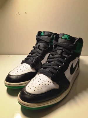 Nike Air Jordan 1 Retro High OG svart/grön - Säljer ett par Nike Air Jordan 1 Retro High OG sneakers i svart, vitt och grönt läder. Klassisk high-top siluett med svart swoosh, gröna detaljer på häl och yttersula samt perforerad tåbox. Perfekta för dig som vill sticka ut med en ikonisk sneaker.