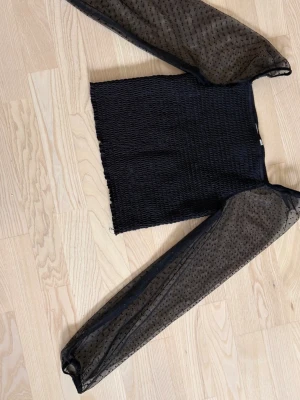 Svart mesh topp med prickiga ärmar - Trendig svart topp med smockad kropp och långa, transparenta meshärmar med små svarta prickar. Perfekt för dig som gillar en edgy och cool stil. Toppen är croppad och har en snygg passform som framhäver midjan.