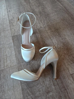 Satinskor med ankelrem st 36 - Satinskor med ankerem ovh remdekor framtill. Färg Ivory Skorna har en tunn ankelrem med guldfärgat spänne och är tillverkade i lyxig satin. Perfekta för dig som vill ha en stilren och klassisk look.