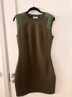 Olivgrön kort klänning med paljetter - Snygg olivgrön kort klänning från NLY ONE med paljettprydda axlar. Ärmlös modell med figurnära passform och rund halsringning. Perfekt för dig som vill sticka ut lite extra. Liten medium