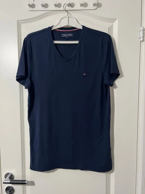 Marinblå v-ringad t-shirt Tommy Hilfiger - Snygg marinblå t-shirt från Tommy Hilfiger med v-ringning och liten broderad logga på bröstet. Tillverkad i mjuk bomull och har korta ärmar. Perfekt basplagg med stilren design. Finns att hämta i Södertälje.