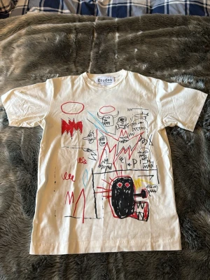 Vit Études t-shirt med konstnärligt tryck - Unik vit t-shirt från Études med färgstarkt, konstnärligt tryck fram och bak i Basquiat-inspirerad stil. T-shirten har korta ärmar, rund hals och är gjord i mjuk bomull. Perfekt för dig som vill sticka ut med en kreativ och cool look. Går ej att köpa längre. 