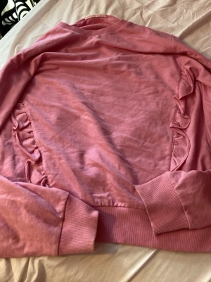 Rosa sweatshirt med volang från KappAhl - Säljer en rosa sweatshirt från KappAhl med volangdetaljer längs sidorna. Tröjan har ribbade muddar vid ärmslut och nederkant samt en klassisk rund halsringning. Perfekt för dig som gillar en söt och avslappnad stil. Aldrig använd 