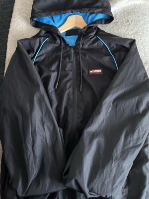 Svart vindjacka från McKenzie - Snygg svart vindjacka från McKenzie med blått meshfoder och blå detaljer vid axlarna. Jackan har huva med dragsko, hel dragkedja framtill och logga på bröstet. Perfekt för blåsiga dagar och enkel att matcha med streetwear. Priset är inte hugget 
