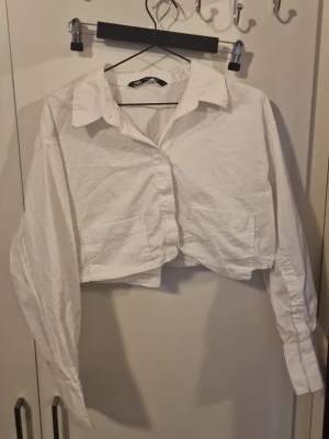 Vit croppad skjorta från Zara - Trendig vit croppad skjorta från Zara med klassisk krage och långa ärmar. Skjortan har knappar framtill och två fickor på bröstet. Perfekt för dig som vill ha en modern twist på en klassisk skjorta.