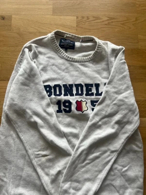 Vit Bondelid tröja med tryck - Vit långärmad tröja från Bondelid med blått tryck 'BONDELID 1955' och en liten sköld i rött och vitt framtill. Rund halsringning och mjukt material, perfekt för en avslappnad stil. Andra bilden så ser man en fläck