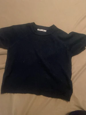 Svart stickad kortärmad tröja - Mysig svart stickad tröja med korta ärmar och rund hals. Tröjan har en enkel och stilren design, perfekt att matcha med olika outfits. Materialet ser ut att vara mjukt och bekvämt, och modellen är något boxig för en avslappnad look.