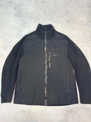 Svart stickad jacka från Moncler - Svart jacka från Moncler med stickade ärmar och hög krage. Jackan har dragkedja framtill, bröstficka med dragkedja och diskret Moncler-logga på bröstet och ärmen. Materialmix med stickad struktur på ärmar och slät front.