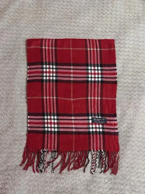 Röd rutig halsduk från Burberrys - Klassisk röd halsduk från Burberrys i 100% lammull med ikoniskt rutmönster i svart, vitt och beige. Halsduken har tvinnade fransar i ändarna och en snygg vävd etikett. Perfekt accessoar för att lyfta din outfit med en tidlös brittisk touch.