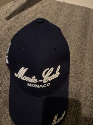 Mörkblå Monte-Carlo keps - Snygg mörkblå keps med vit brodyr där det står 'Monte-Carlo Monaco' framtill. Klassisk böjd skärm och justerbar baksida. Perfekt accessoar för en sportig och trendig look.