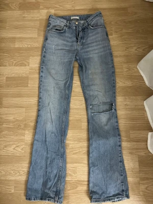 Ljusblå bootcut jeans med slitning - Säljer ett par ljusblå bootcut jeans från Gina tricot med rå kant och en tydlig slitning på ena benet. 