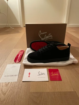 Svarta sneakers från Christian Louboutin - Snygga svarta sneakers från Christian Louboutin i mocka med klassisk röd sula. Lågt skaft, svarta skosnören och diskret design som ändå sticker ut tack vare den ikoniska röda undersidan. Perfekta för dig som vill ha något exklusivt och stilrent.