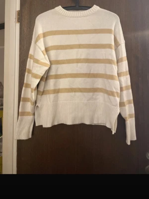 Beige och vit randig stickad tröja - Säljer en oversized stickad tröja med breda beige och vita ränder. Tröjan har rund halsringning, långa ärmar och ribbade muddar samt nederkant. Perfekt för dig som gillar en avslappnad och trendig stil.